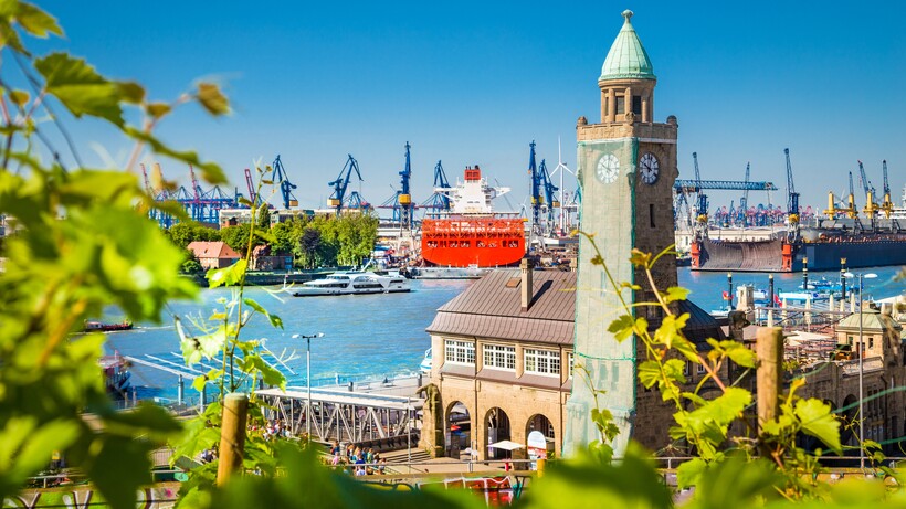 Farbfoto mit Blick Landungsbrücken und Uhrenturm bei blauem Himmel, im Hintergrund Hafenanlage.